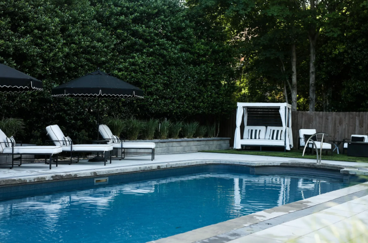 Poolside scene with pool, lounge chairs, black umbrellas, and white cabana. Green hedges and trees in the background.