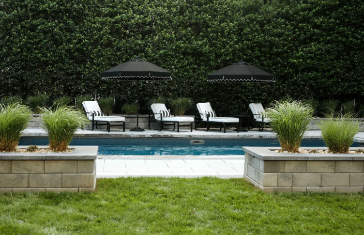Lounge chairs and black umbrellas by a pool, framed by hedges and stone planters with ornamental grass.