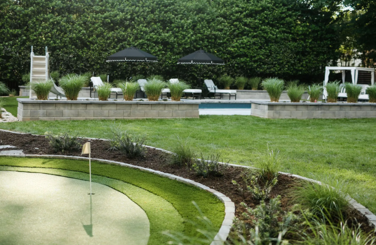 Backyard with a putting green, pool, and hedge. Lawn chairs and umbrellas near the pool.