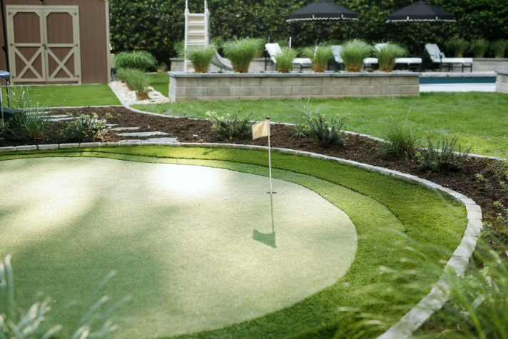 Backyard putting green with flag, grass, and stone border; shed and pool in background.