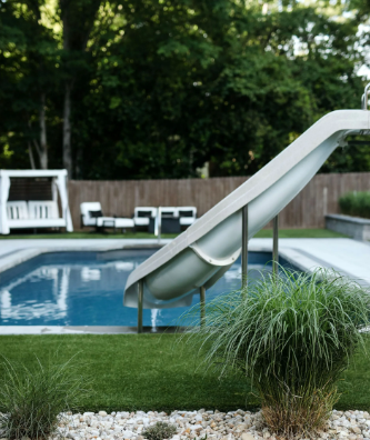 Pool with slide, lounge chairs, and a swing set in a backyard setting with green grass.