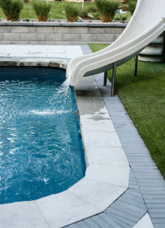 Pool with white slide. Water flows from slide into blue water. Light gray stone edging. Green grass nearby.