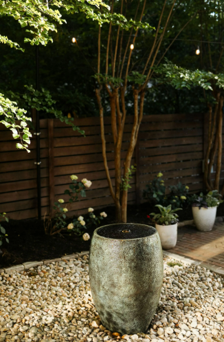 A weathered, stone water fountain in a gravel patio, with potted plants, a wooden fence, and trees.