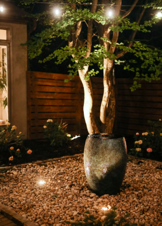 Tree in decorative pot, lit by string lights and spotlights, set in a gravel garden at night.
