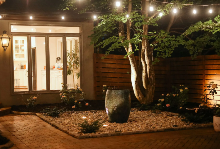 Backyard patio with string lights, a water fountain, and a tree at night.