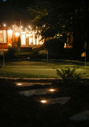 Nighttime scene of a lit golf green with flags, a stream, and a building with string lights in the background.