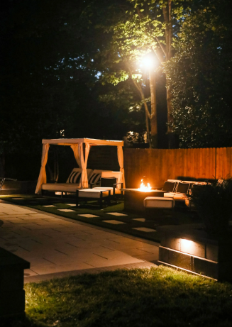 Nighttime backyard scene with lit fire pit, gazebo with cushions, and path lit by a lamp.