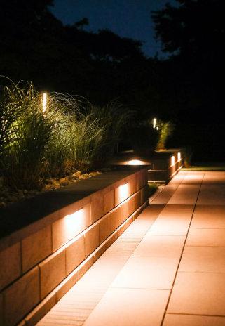 Pathway lit by warm lights along a stone wall and bordering plants at night.
