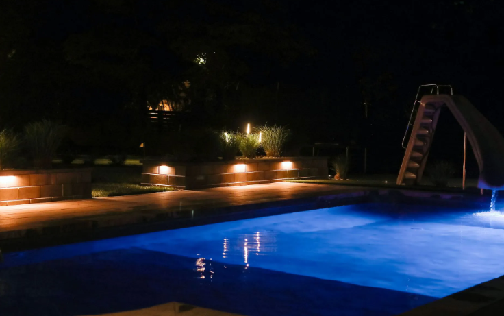 Pool illuminated at night with a slide and surrounding lights reflecting in the blue water.
