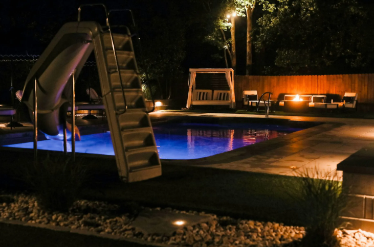 Pool area lit at night, pool slide, lounge furniture, small lights, trees, and a wooden fence.