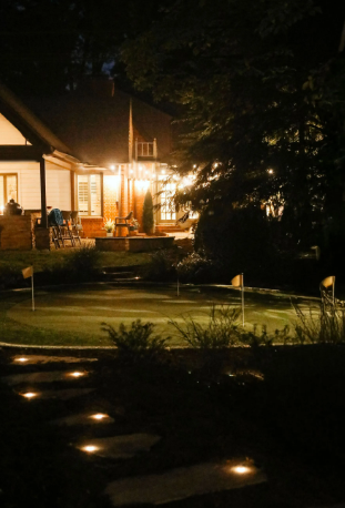 Nighttime view of a backyard with a putting green, lit steps, and a house with warm lights.