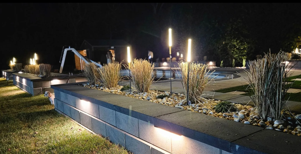 Nighttime landscape with illuminated retaining walls, tall lights, and ornamental grasses.