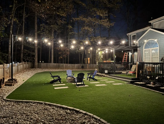 Backyard at night with string lights, fire pit, seating, and house. Green grass with stone pathways.