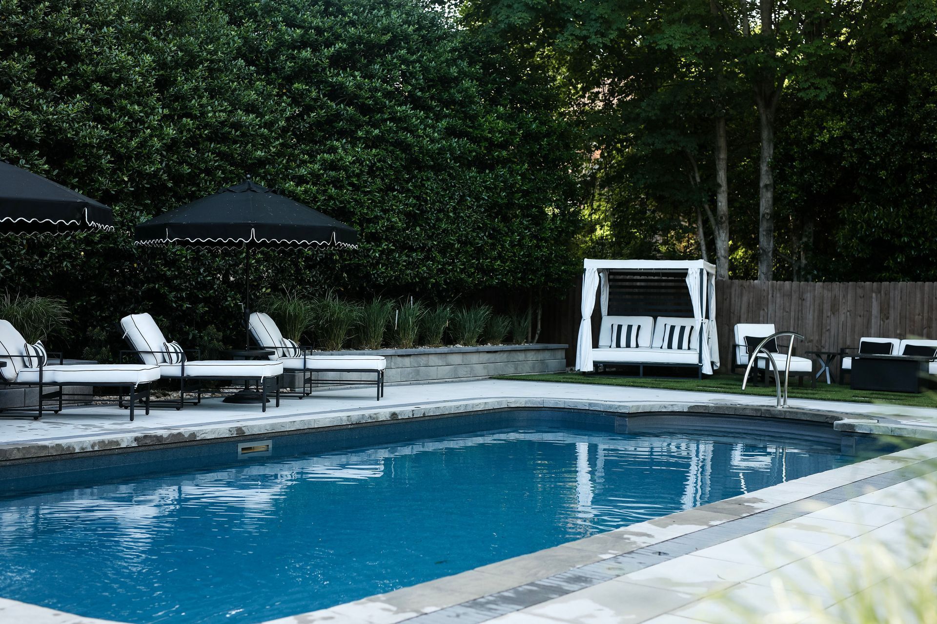 Empty swimming pool with beige stone decking and a white house in the background. Blue tile trim.