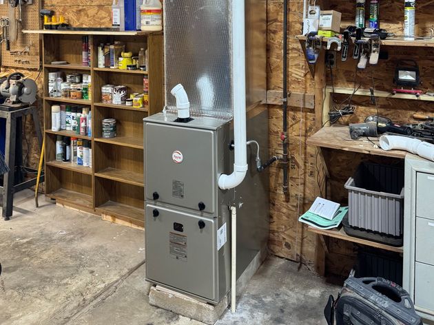 Workshop with metal furnace, white ducting, shelves of supplies, and a cluttered workbench.