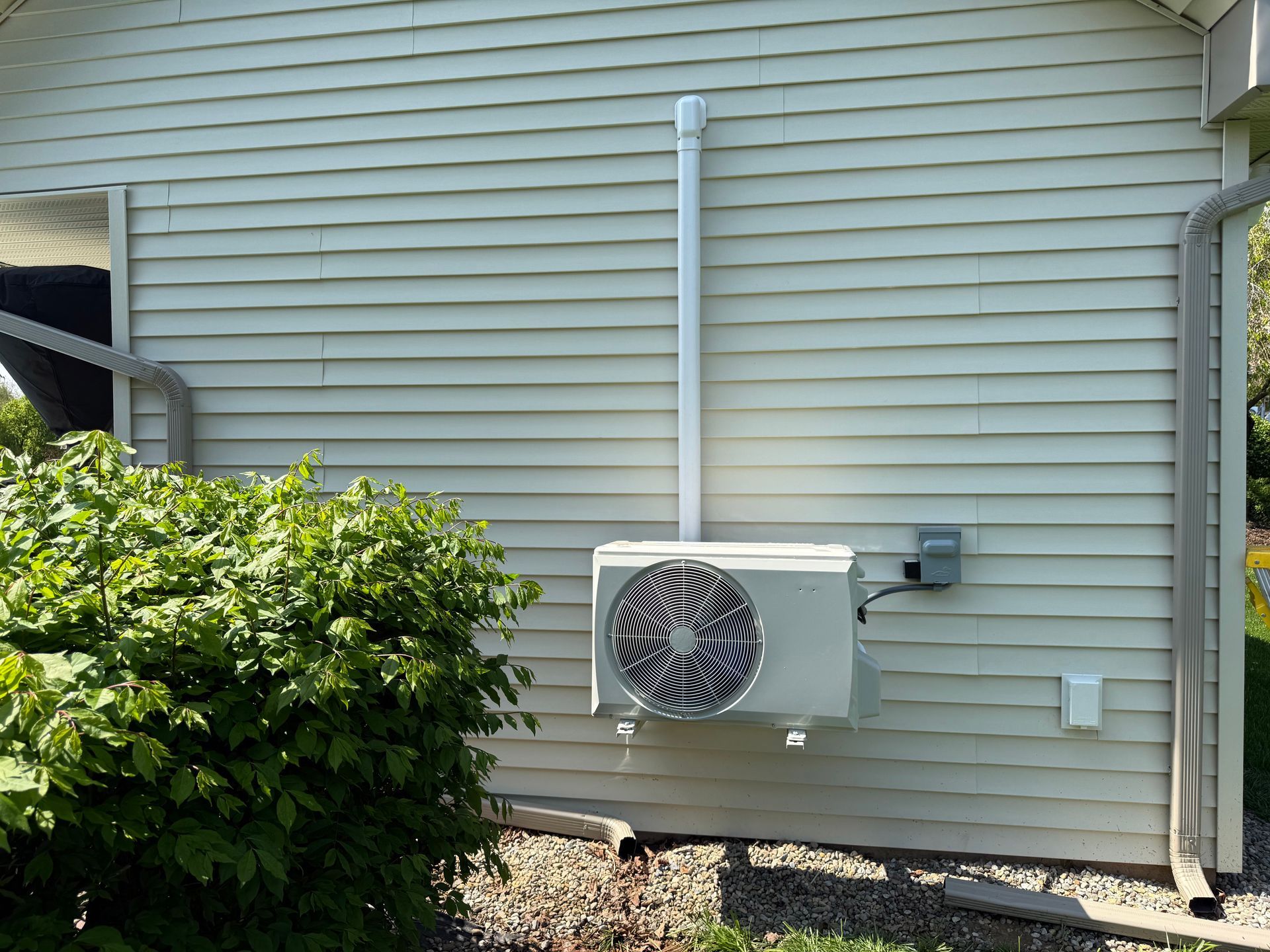 Outdoor air conditioning unit mounted on beige house siding beside green bushes