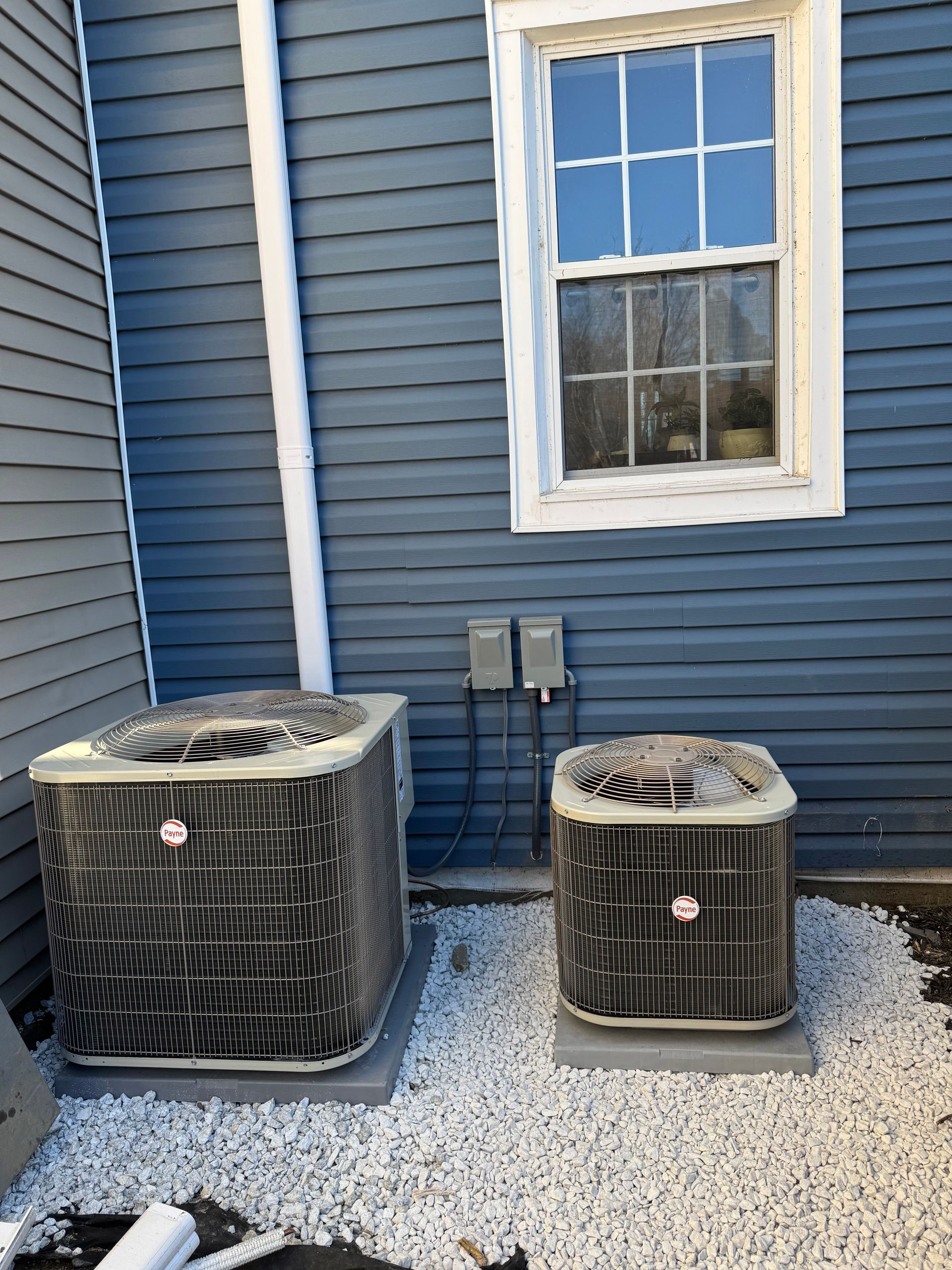 Two outdoor AC units beside a blue house wall under a window, with snow on the ground.