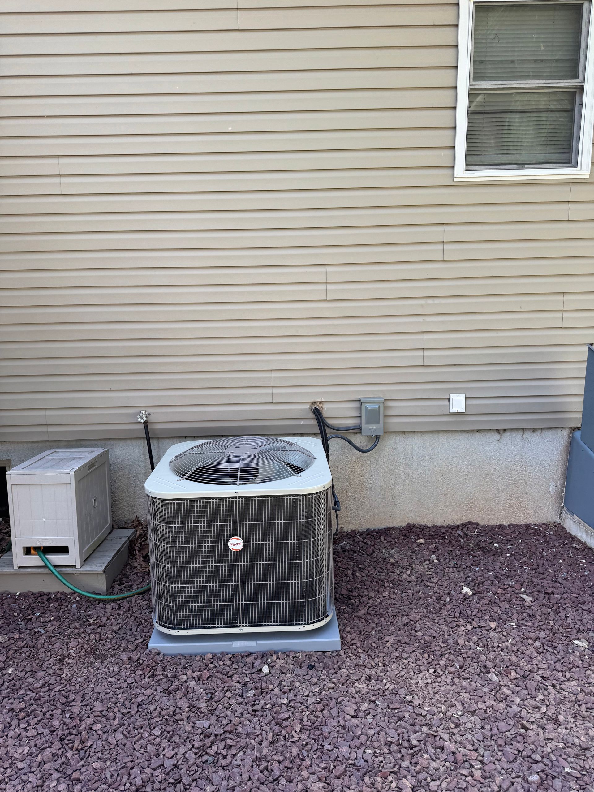 Outdoor HVAC unit beside a beige house wall with a window and utility box on gravel.