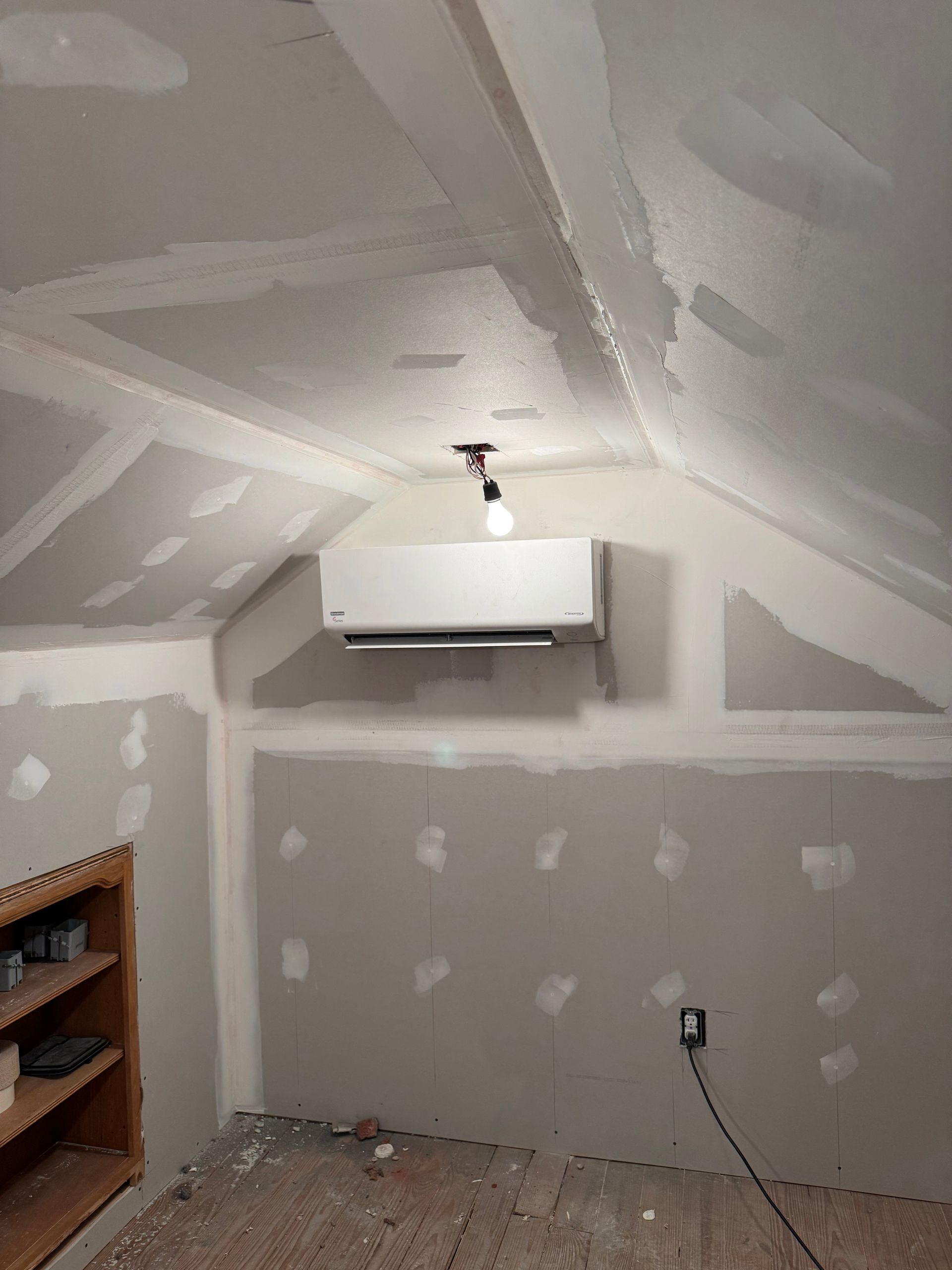 Unfinished attic room with sloped ceiling, white and gray drywall, wall-mounted AC unit, and a bare light bulb.