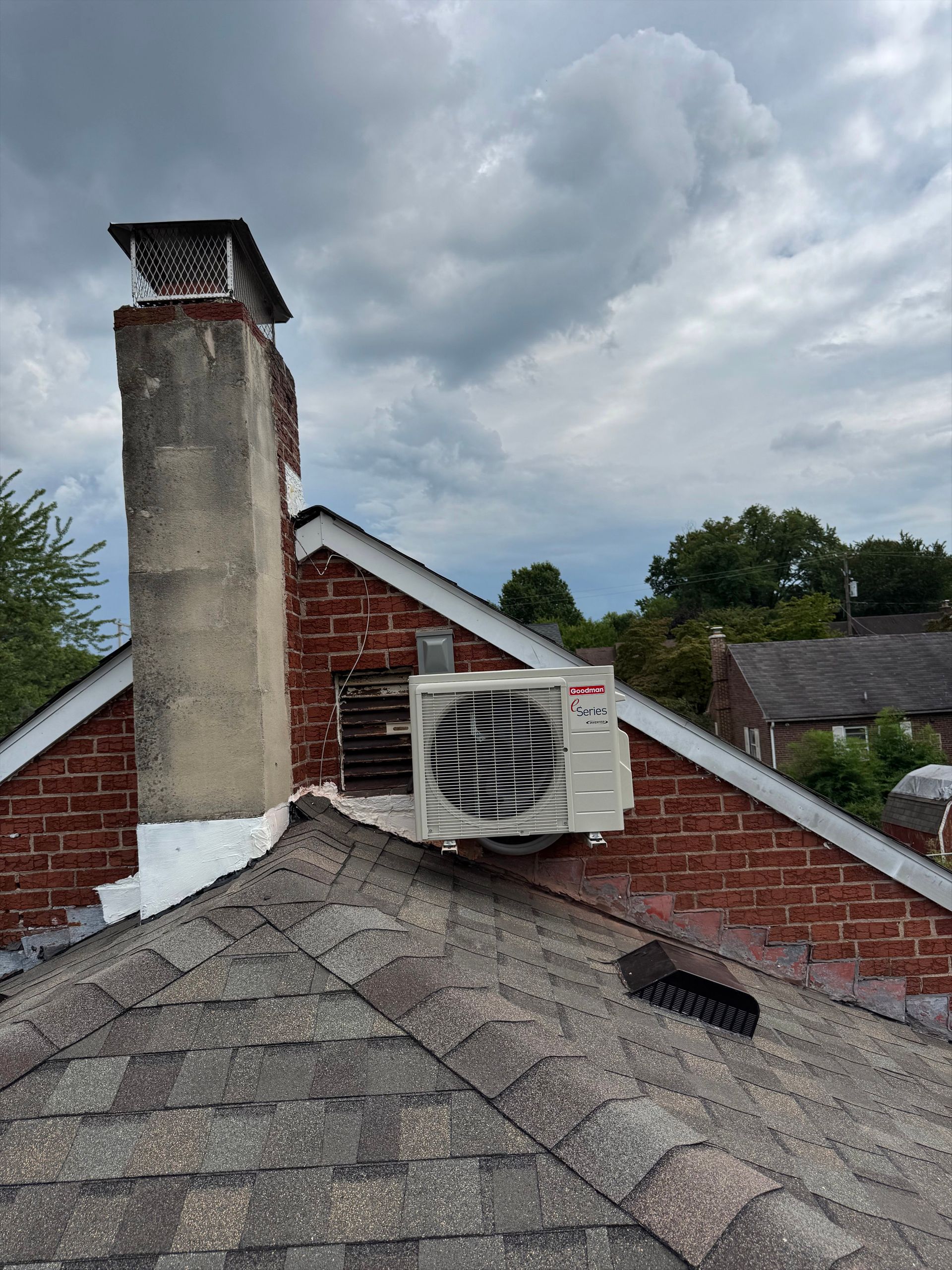 Rooftop vent unit beside a brick chimney on a shingled roof under cloudy skies