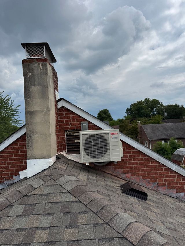 Rooftop vent unit beside a brick chimney on a shingled roof under cloudy skies