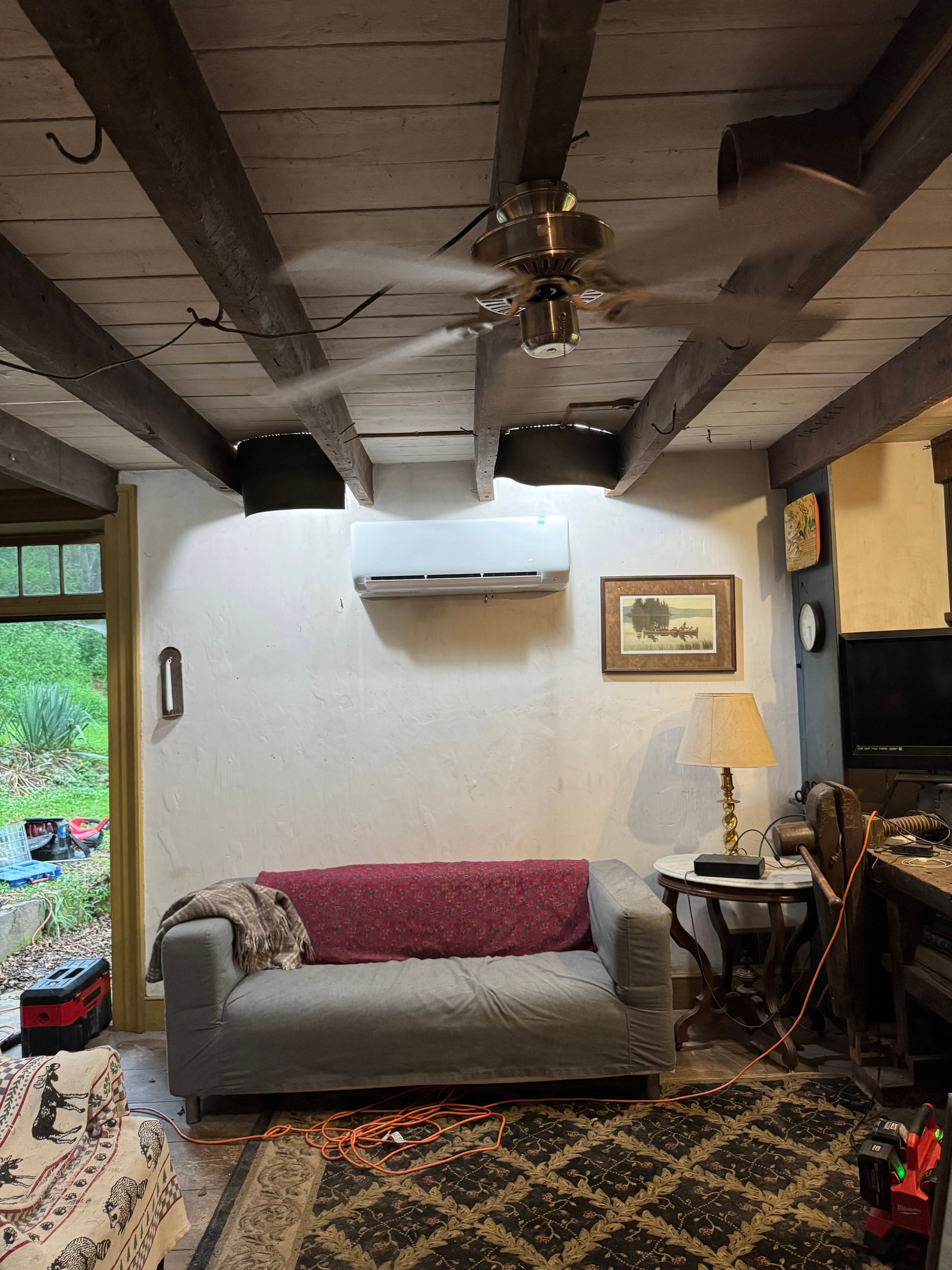 Rustic living room with exposed wood beams, gray sofa, ceiling fan, wall AC, lamp, and patterned rug.