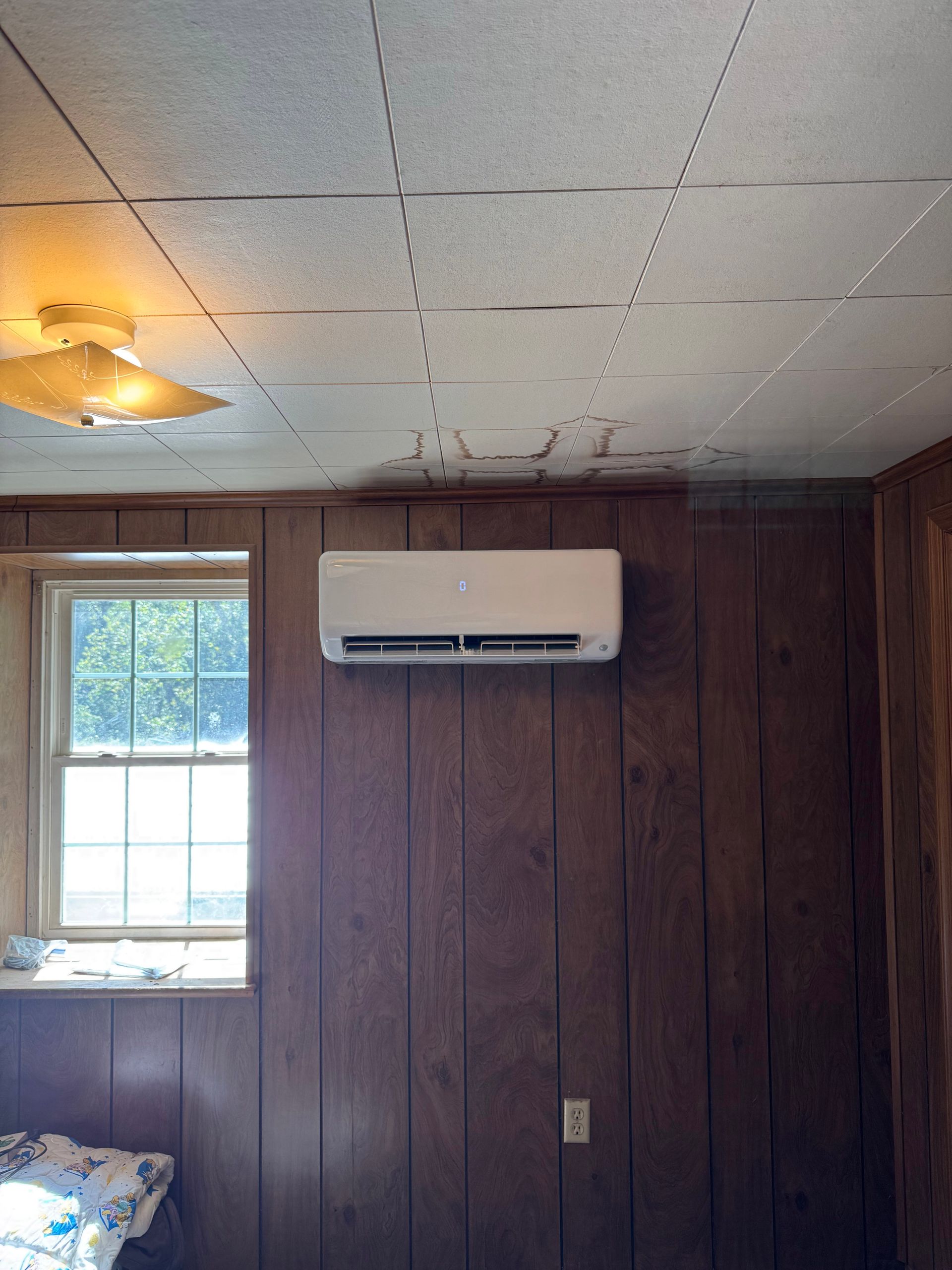 Small room with wood-paneled wall, window, ceiling fan light, and wall-mounted air conditioner.