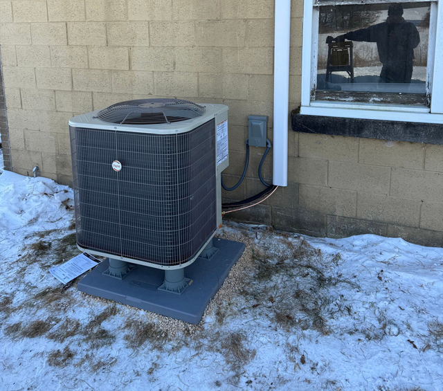 Outdoor air conditioner unit beside a tan brick wall with snow on the ground and a window nearby
