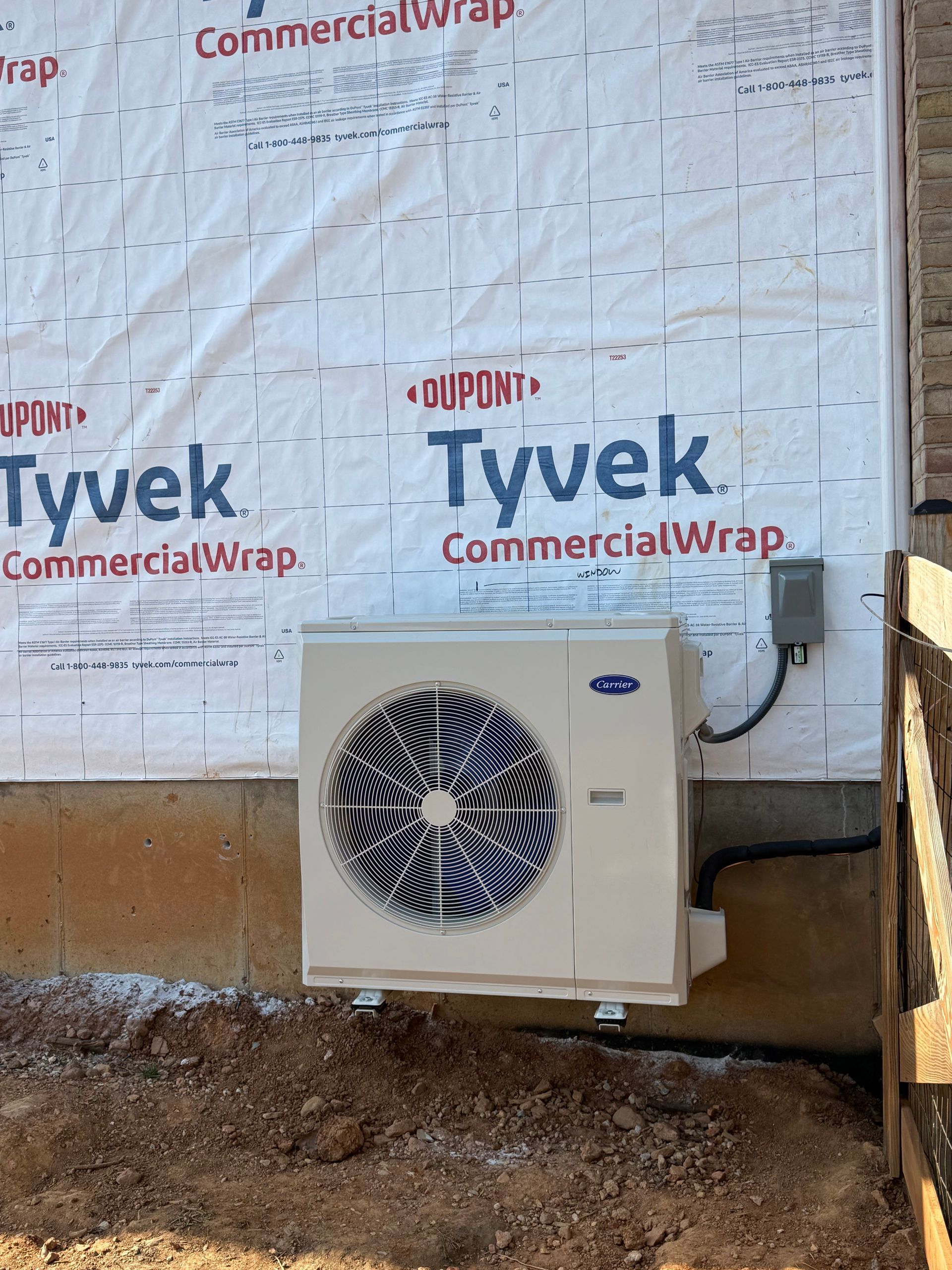Outdoor HVAC condenser unit beside a house wrapped in Tyvek, sitting on a dirt foundation.