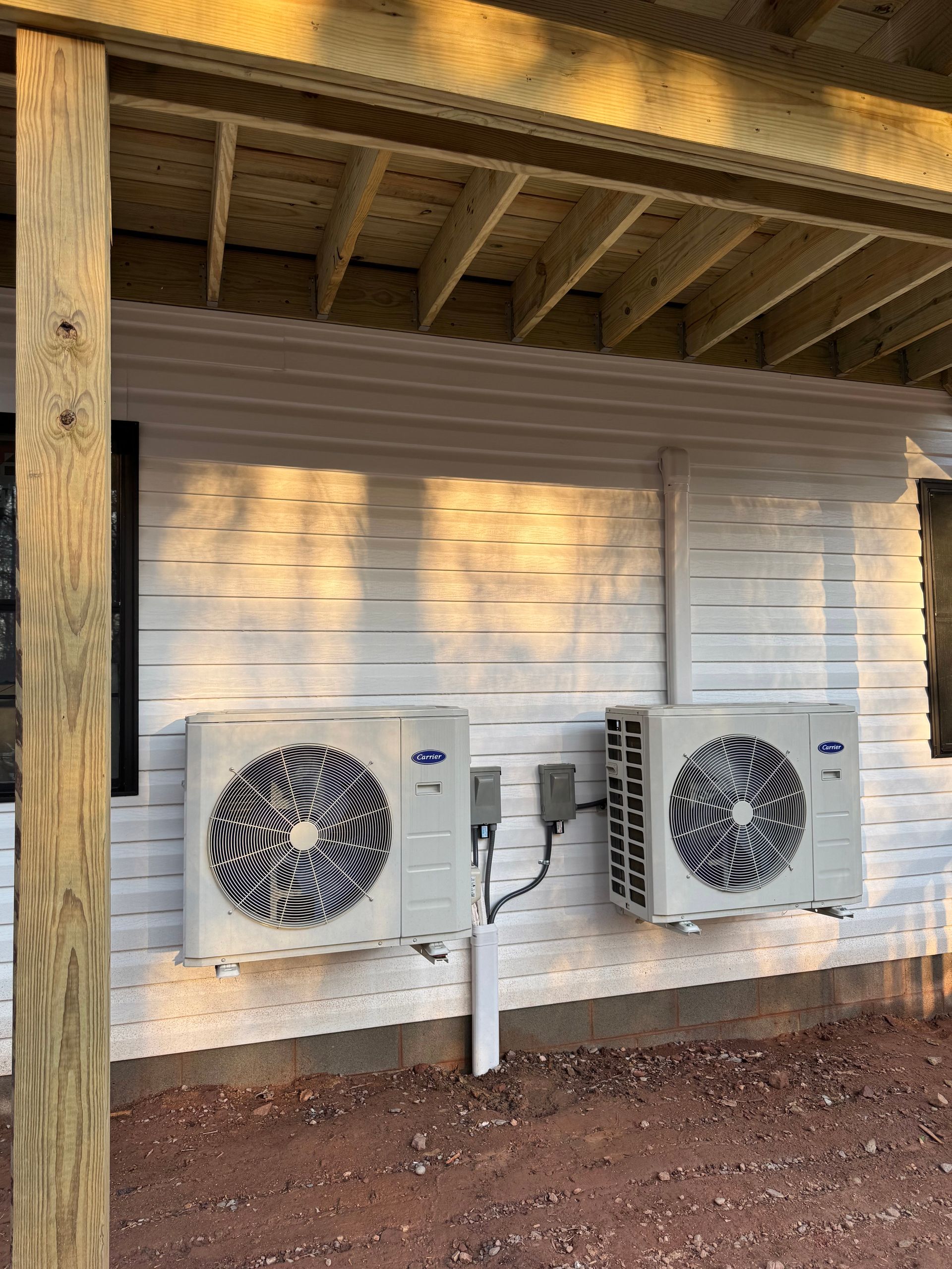 Two outdoor HVAC units mounted on a white wall under a wooden porch roof.