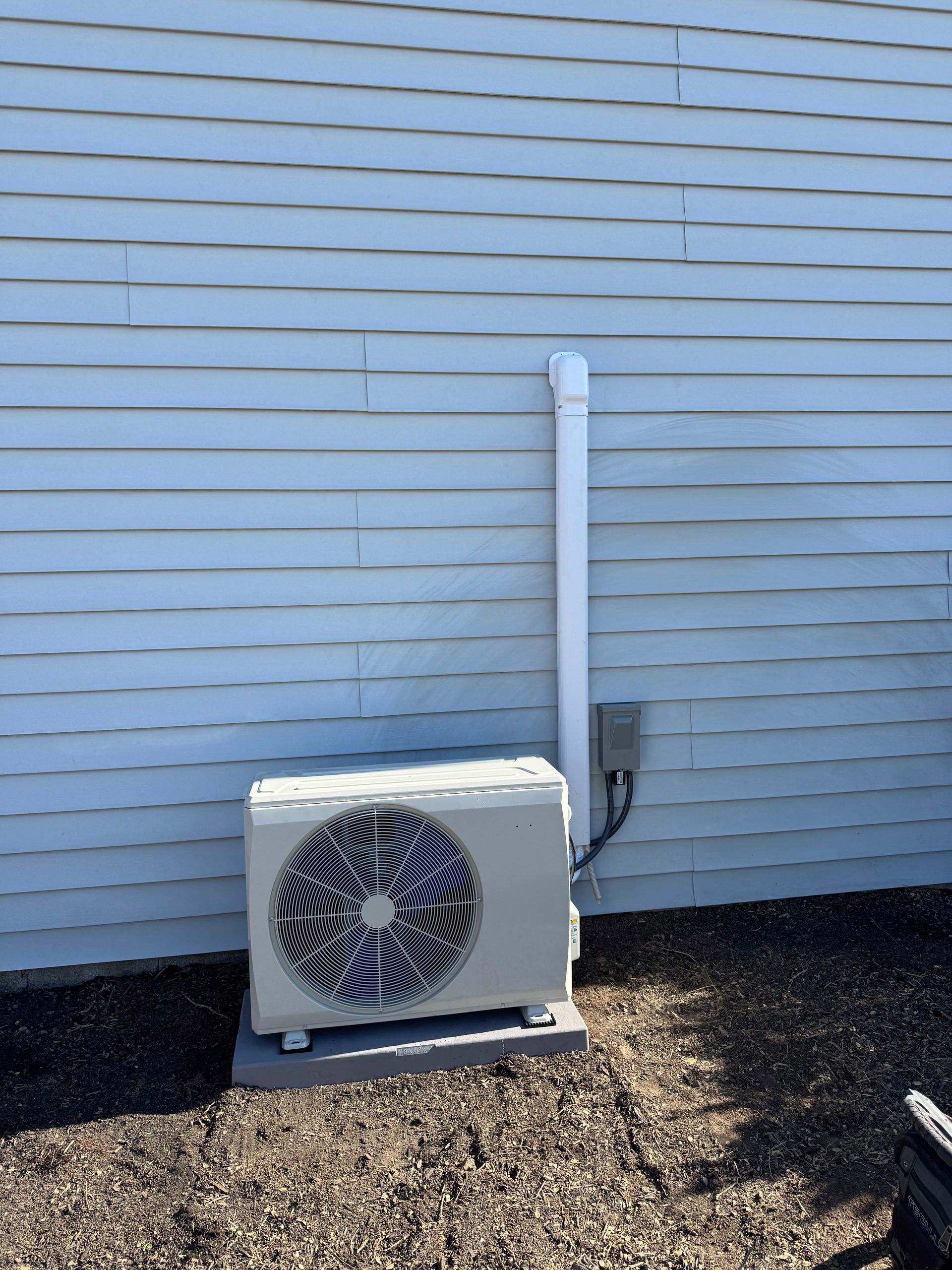 Outdoor HVAC unit beside a white siding wall with a vertical white pipe and gravel ground