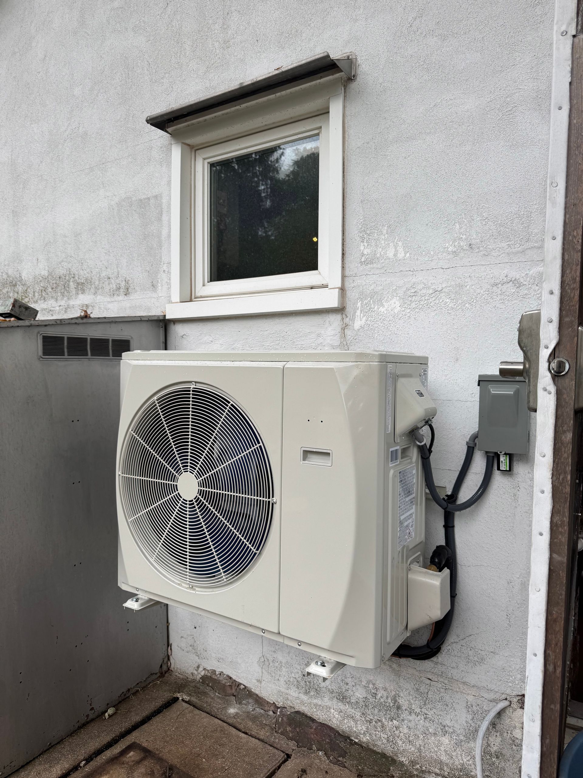 Outdoor wall-mounted air conditioner unit beneath a window on a gray stucco wall.