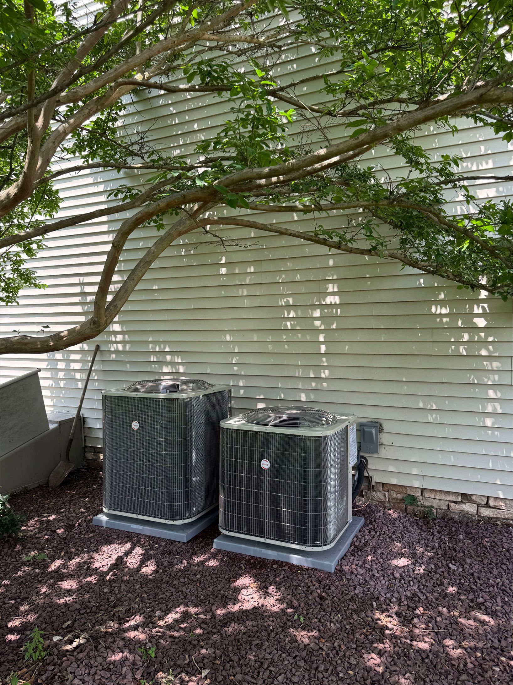 Two outdoor air conditioning units beside a white house under a leafy tree