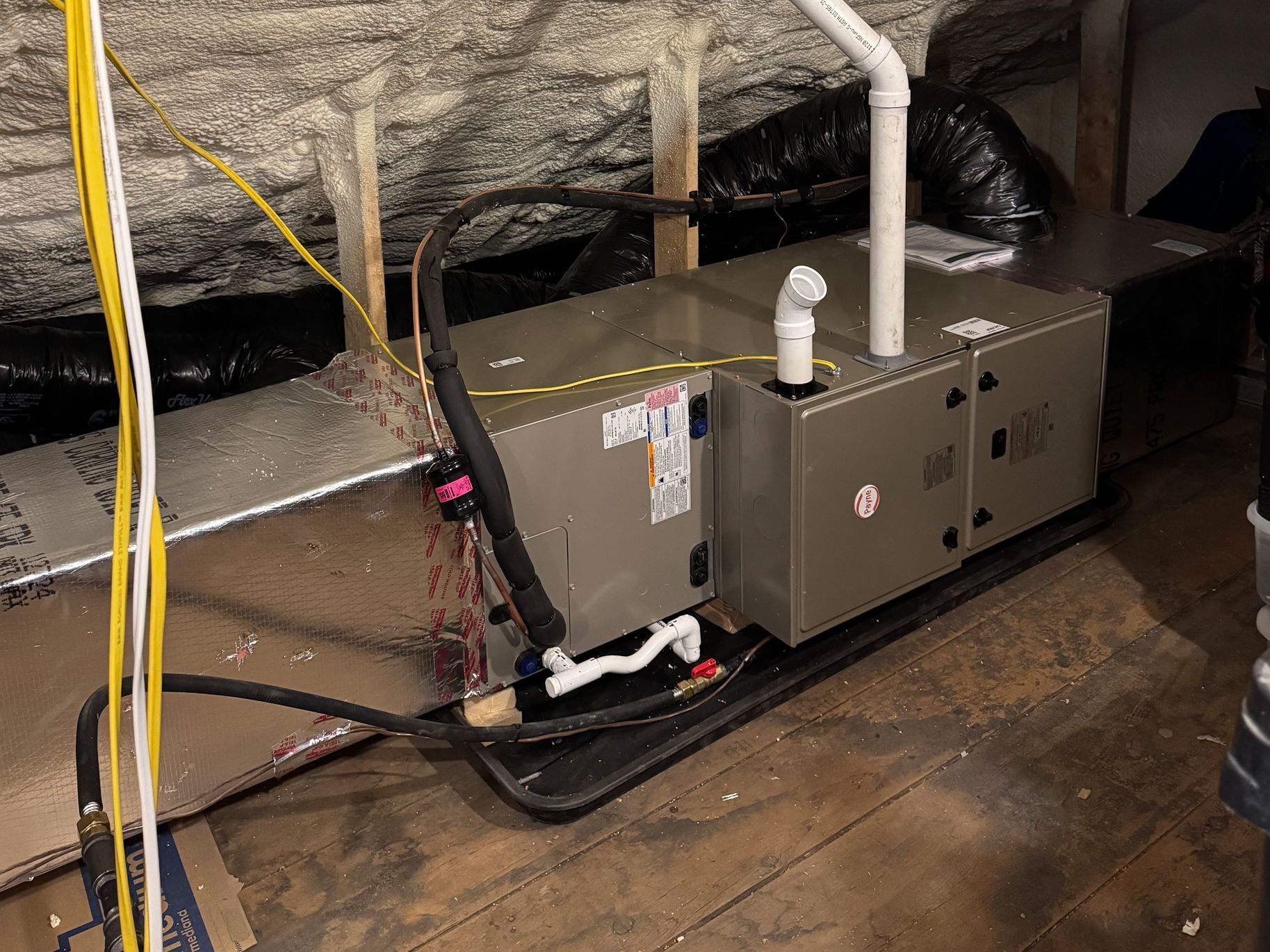 Gray HVAC unit in a crawl space with white pipes and yellow wires under reflective insulation.