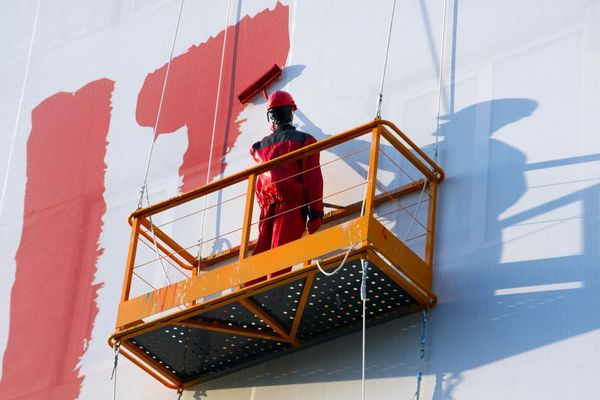 Worker on orange lift painting a white wall red outdoors