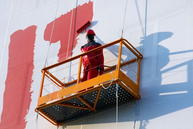 Worker on orange lift painting a white wall red outdoors