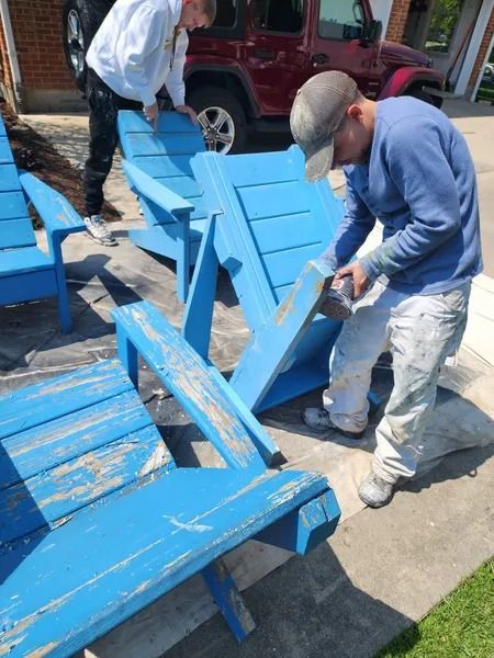 Two people repainting blue wooden chairs on a sidewalk near a parked car