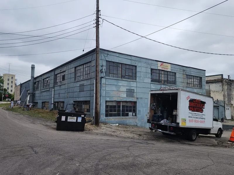 Dilapidated blue warehouse with a white truck parked beside a dumpster on a gravel lot.