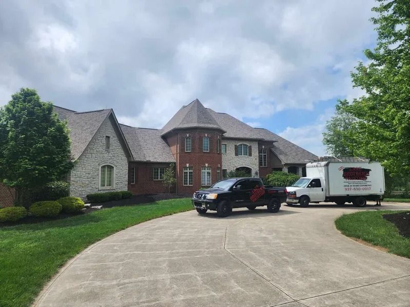 Large brick house with a curved driveway, parked black truck and white trailer under a cloudy sky
