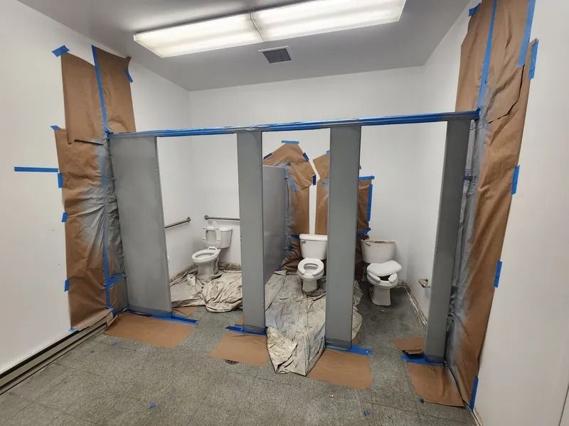 Bathroom under renovation with three toilet stalls, blue tape, and plastic-covered walls and fixtures