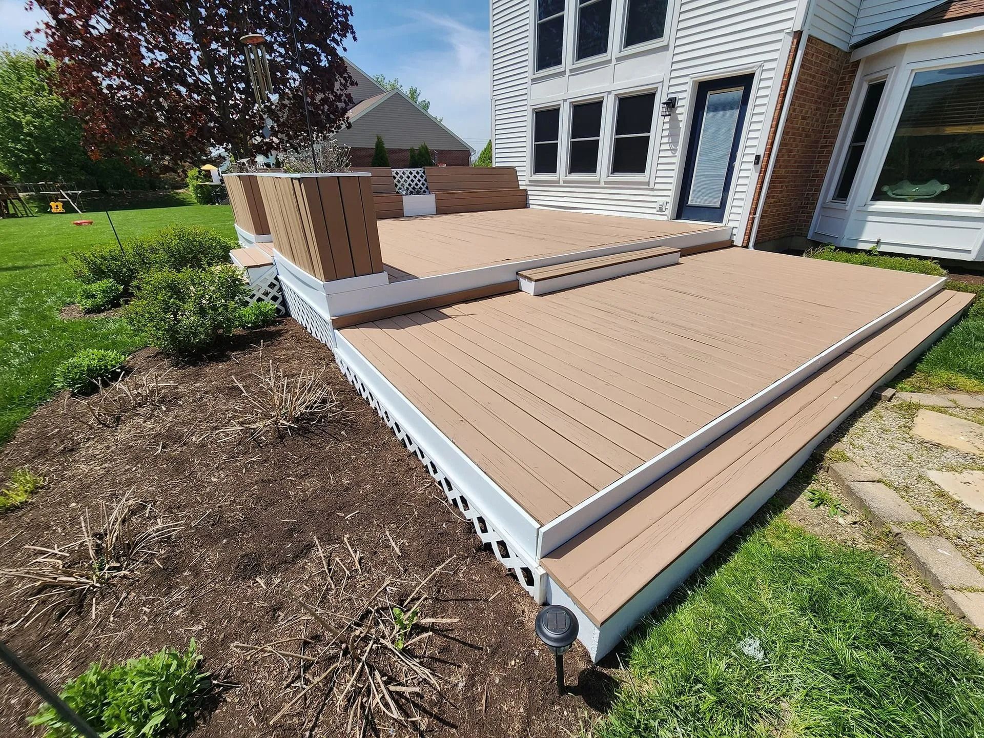 Raised composite deck with stairs beside a house, featuring tan boards and white trim.