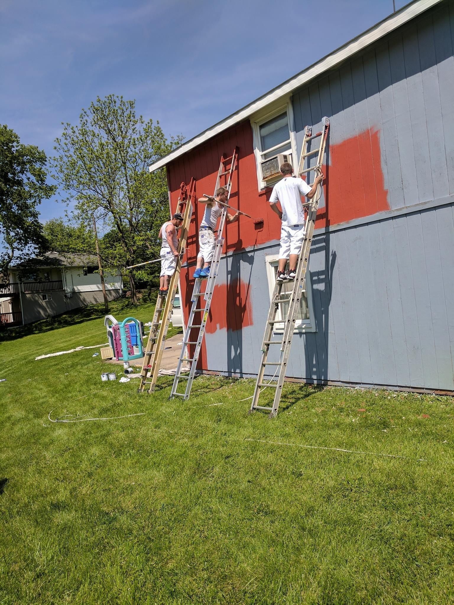 People on ladders painting a gray building red and white on a sunny grassy lawn