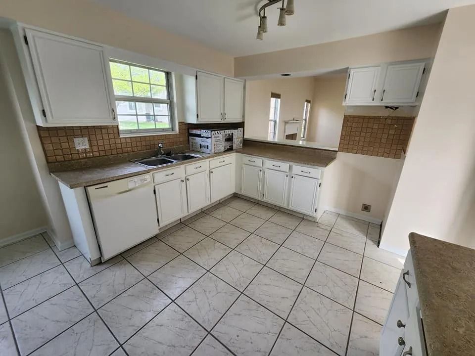 Empty kitchen with white cabinets, tan tile backsplash, and large tiled floor, viewed from the corner.