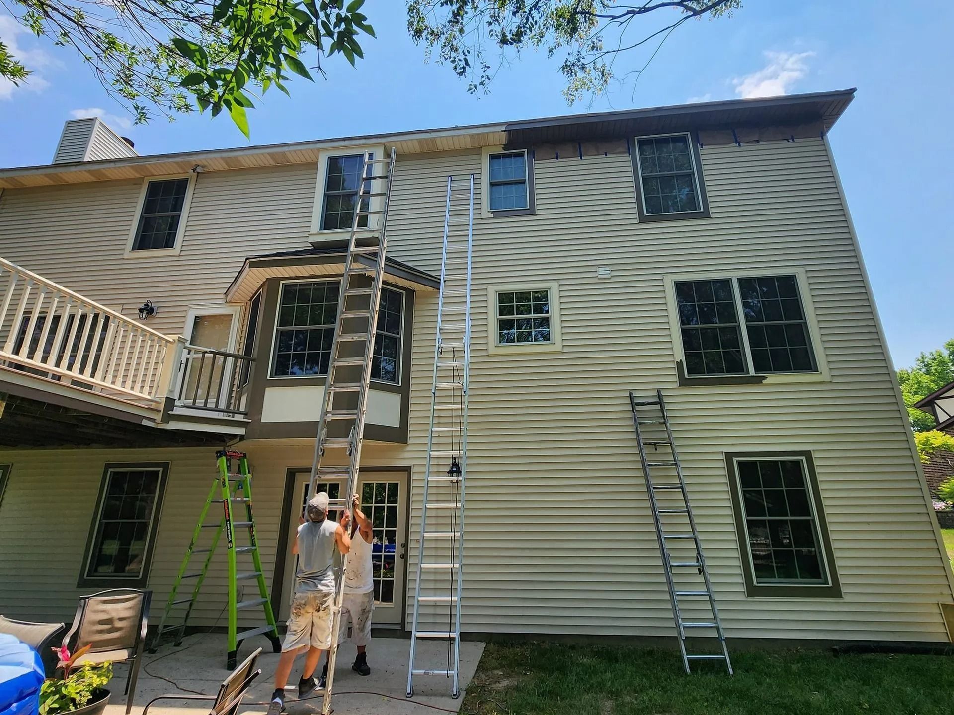 Two workers on ladders painting a two-story beige house exterior on a sunny day