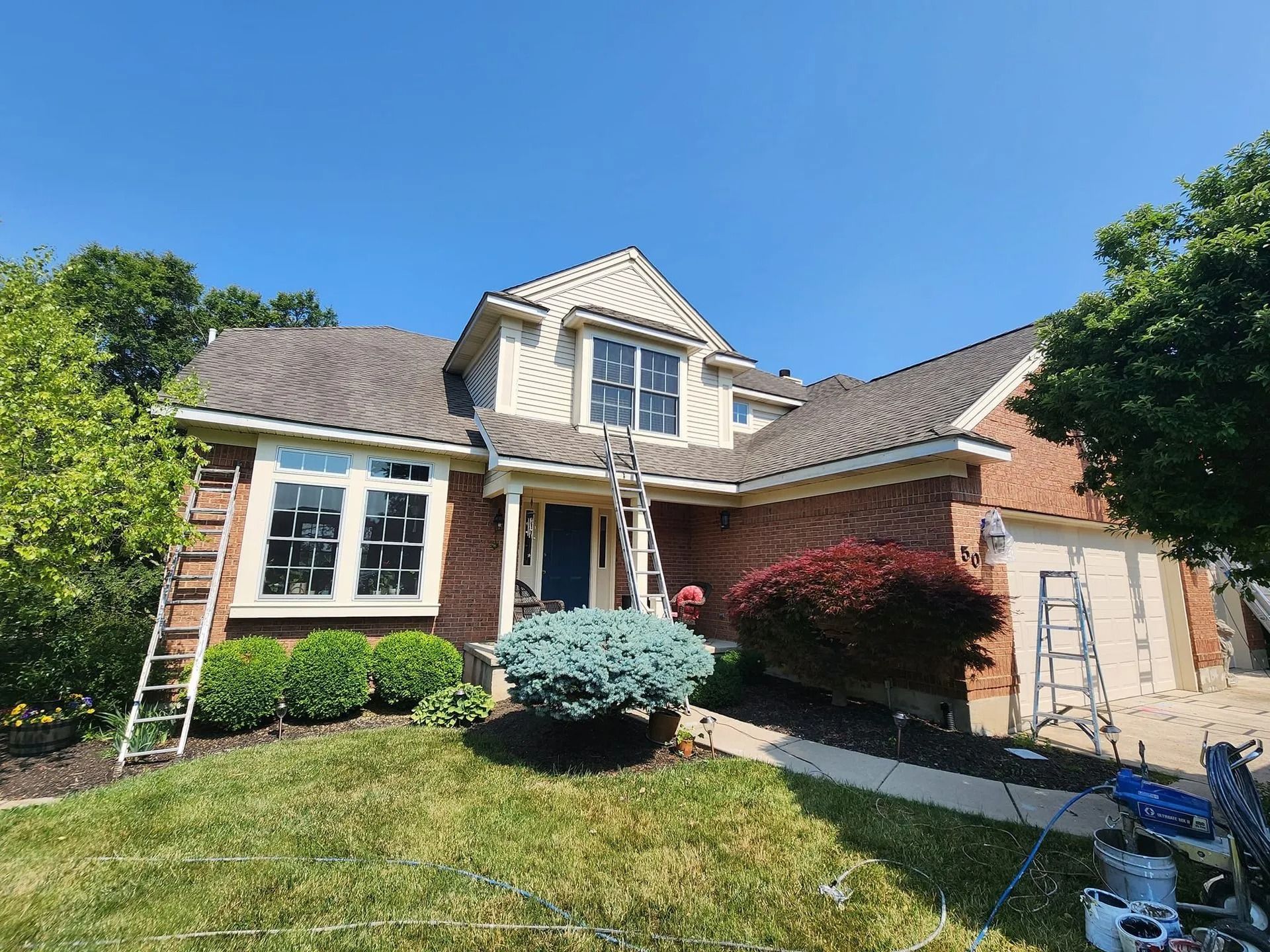 Front view of a brick house with blue sky, ladders on the lawn, and bushes by the porch