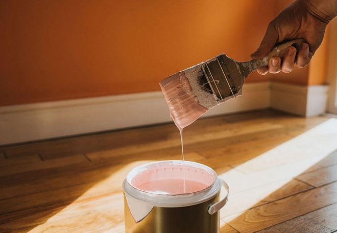 Hand holding a paintbrush dripping pink paint into a paint can on a wooden floor