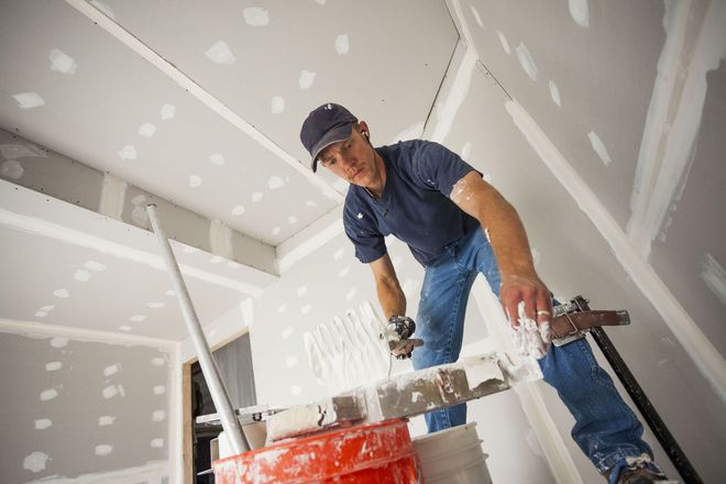 Worker taping drywall seams in an unfinished room with a ladder and tools