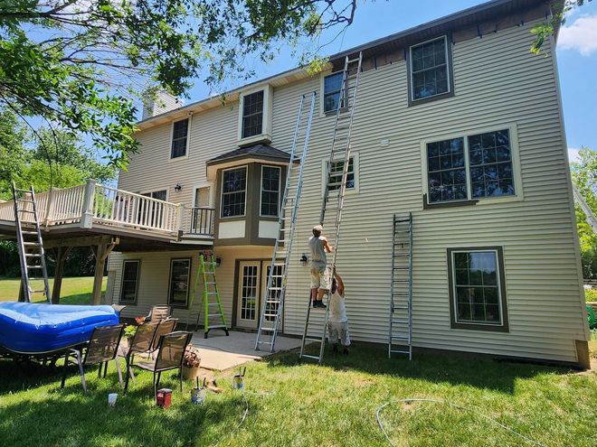 Workers on ladders painting the exterior of a two-story house with a deck and yard.