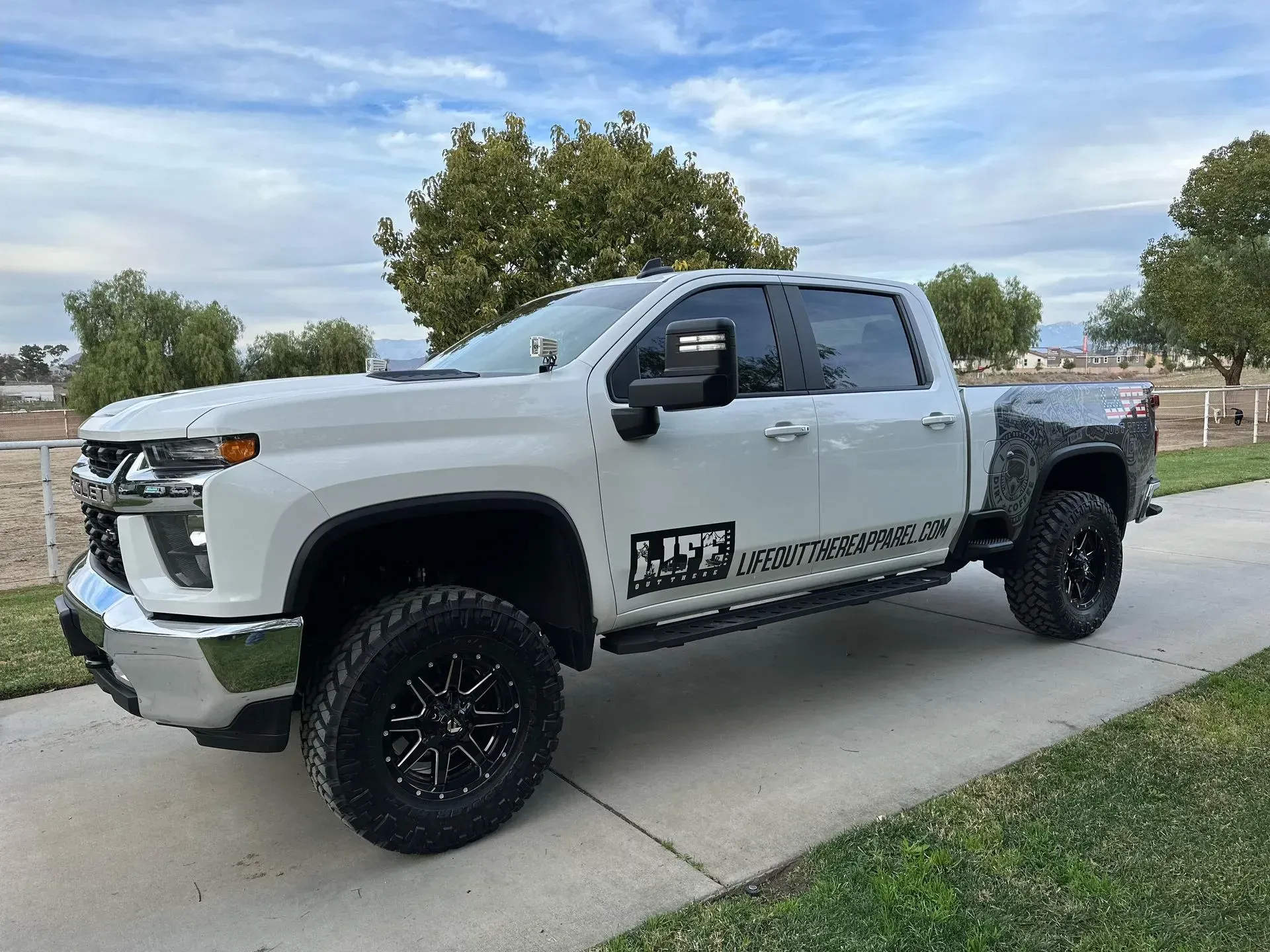 White Chevrolet Silverado truck with black rims on a paved driveway, parked on a grassy area, overcast sky in the background.