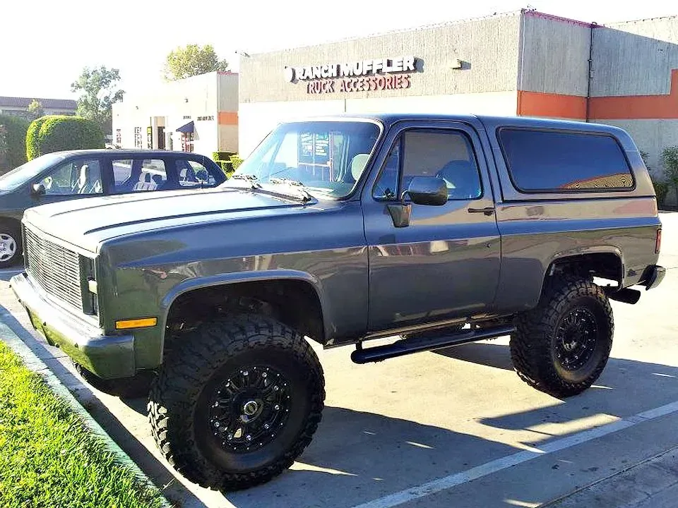 Gray lifted SUV with black wheels parked in a lot, building background.
